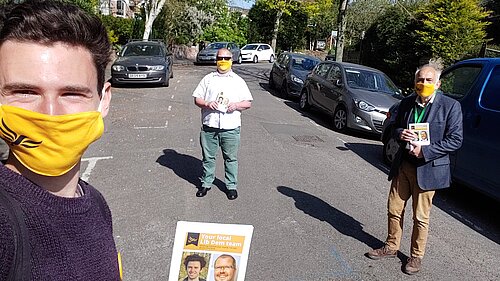 Three Exeter Lib Dems stand, physically-distanced and with masks on, on Prospect Park. The one closest to the camera holds up a canvassing card.