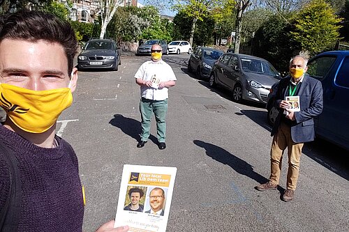 Three Exeter Lib Dems stand, physically-distanced and with masks on, on Prospect Park. The one closest to the camera holds up a canvassing card.