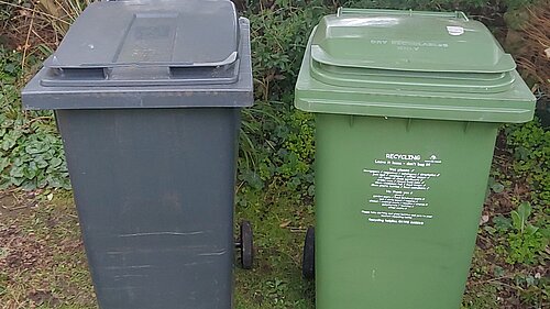 A black bin on the left and a green recycling bin on the right stand in a garden. A service provided by Exeter City Council.