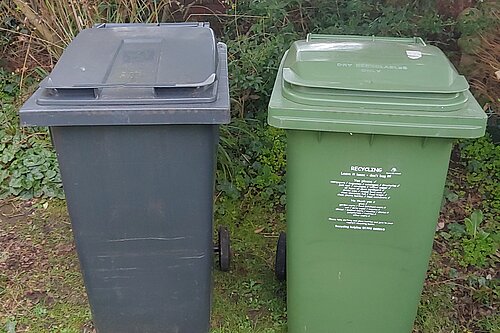 A black bin on the left and a green recycling bin on the right stand in a garden. A service provided by Exeter City Council.