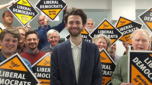 Will Aczel in front of a crowd of Exeter Lib Dems holding up Liberal Democrats working for you posterboards