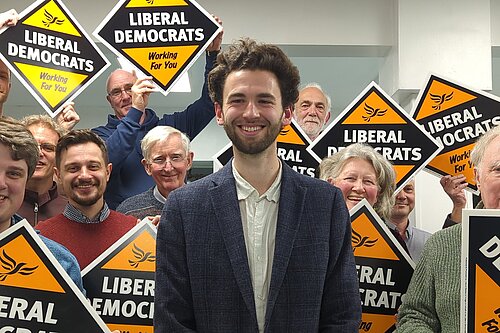Will Aczel in front of a crowd of Exeter Lib Dems holding up Liberal Democrats working for you posterboards