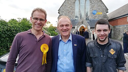 Adrian Fullam, Ed Davey, and Jamie Horner smile at the camera in front of a church in Honiton after a rally for Richard Foord in the Tiverton & Honiton by-election