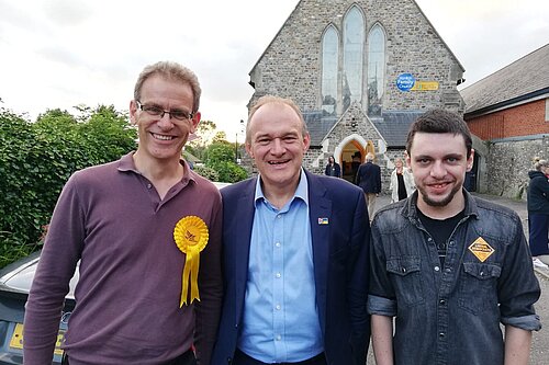 Adrian Fullam, Ed Davey, and Jamie Horner smile at the camera in front of a church in Honiton after a rally for Richard Foord in the Tiverton & Honiton by-election