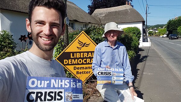 Will Aczel and Charles hold up Our NHS is in Crisis leaflets in front of a Lib Dem Demand Better posterboard